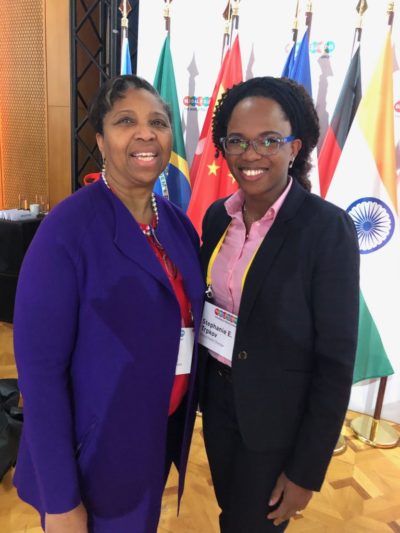 Two women smile together in front of flags from various countries, showcasing a friendly and professional atmosphere.
