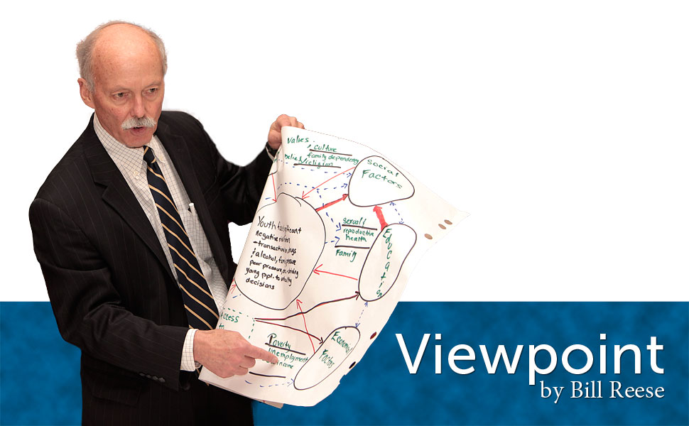 A man in a suit holds a large paper with written content and diagrams, pointing to it while standing against a blue background.