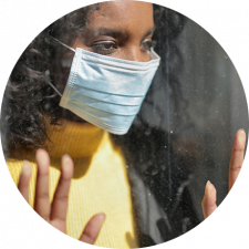 A woman wearing a mask gazes out a window, pressing her hands against the glass, conveying a sense of longing or isolation.