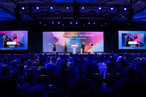A stage with speakers presenting at a large conference, illuminated by blue lights, with an audience in the foreground.