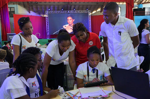 A group collaboration among students at a conference, focused on a laptop, with a speaker visible on a large screen in the background.