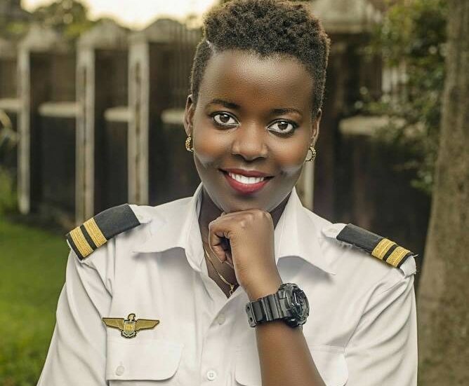 A smiling female pilot in uniform poses confidently, showcasing her watch against a green, outdoor background.