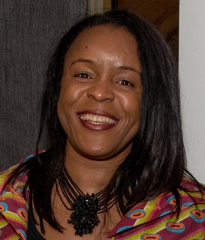 A smiling woman with long, dark hair, wearing a colorful patterned garment and a statement necklace.
