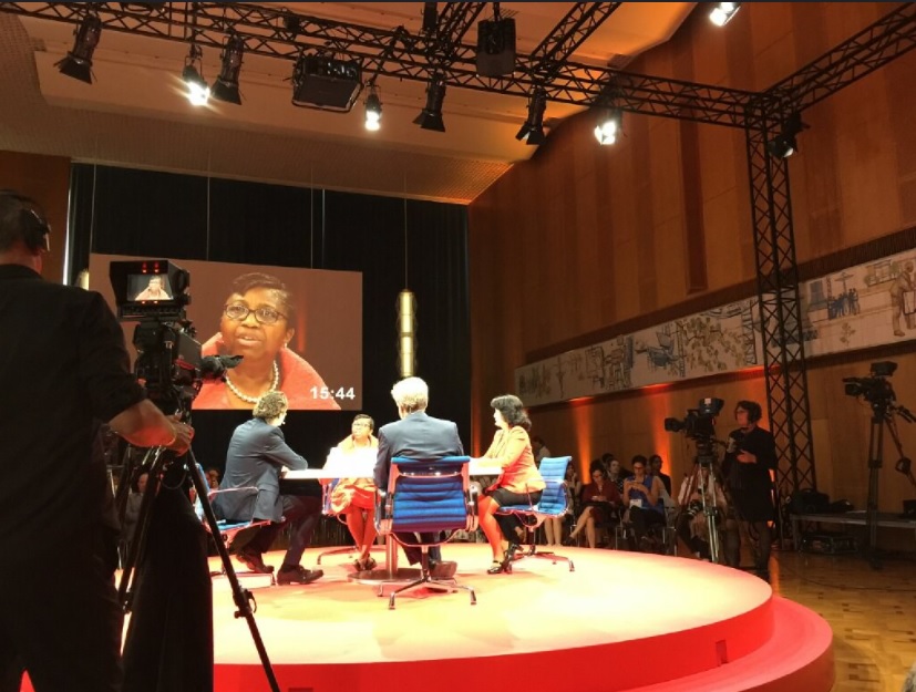 A panel discussion on a red circular stage with five speakers and an audience in a well-lit auditorium.