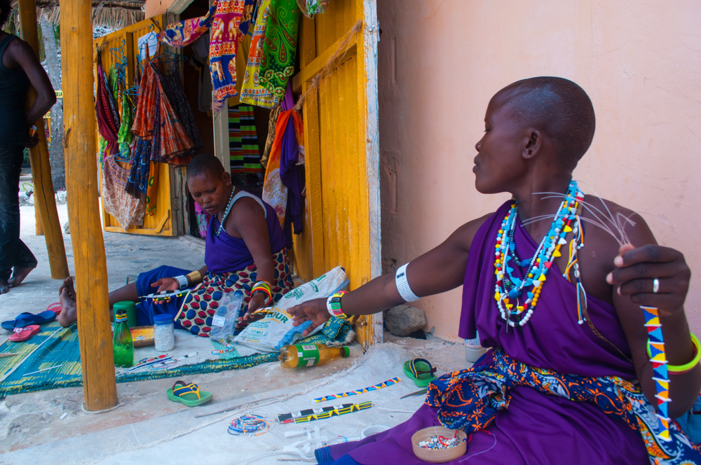Two women adorned in traditional attire craft jewelry surrounded by colorful textiles in a vibrant outdoor setting.