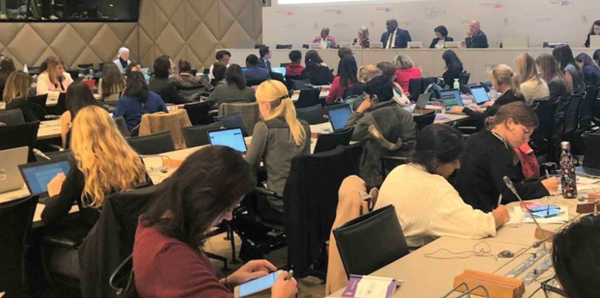 A large group of people engaged in a conference, with many using laptops and taking notes in a well-lit meeting room.