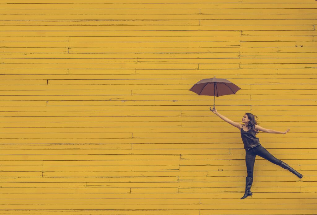 A woman playfully poses with a brown umbrella, balancing on one leg against a vibrant yellow wooden wall.