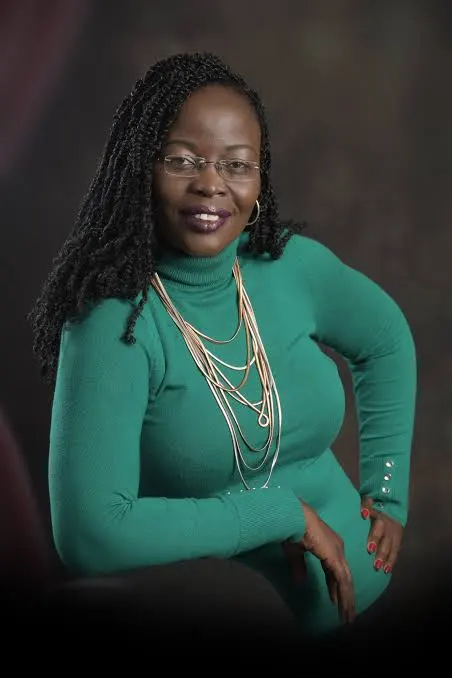 A woman in a green turtleneck dress poses gracefully, wearing layered necklaces, with a confident smile against a blurred background.