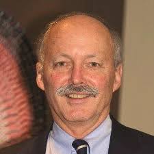 A middle-aged man with a mustache, dressed in a suit, smiles at the camera against a blurred background.
