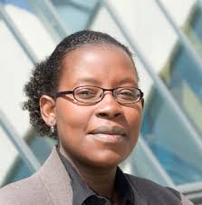 A professional woman with glasses, wearing a dark blazer, stands outdoors with a modern building in the background.