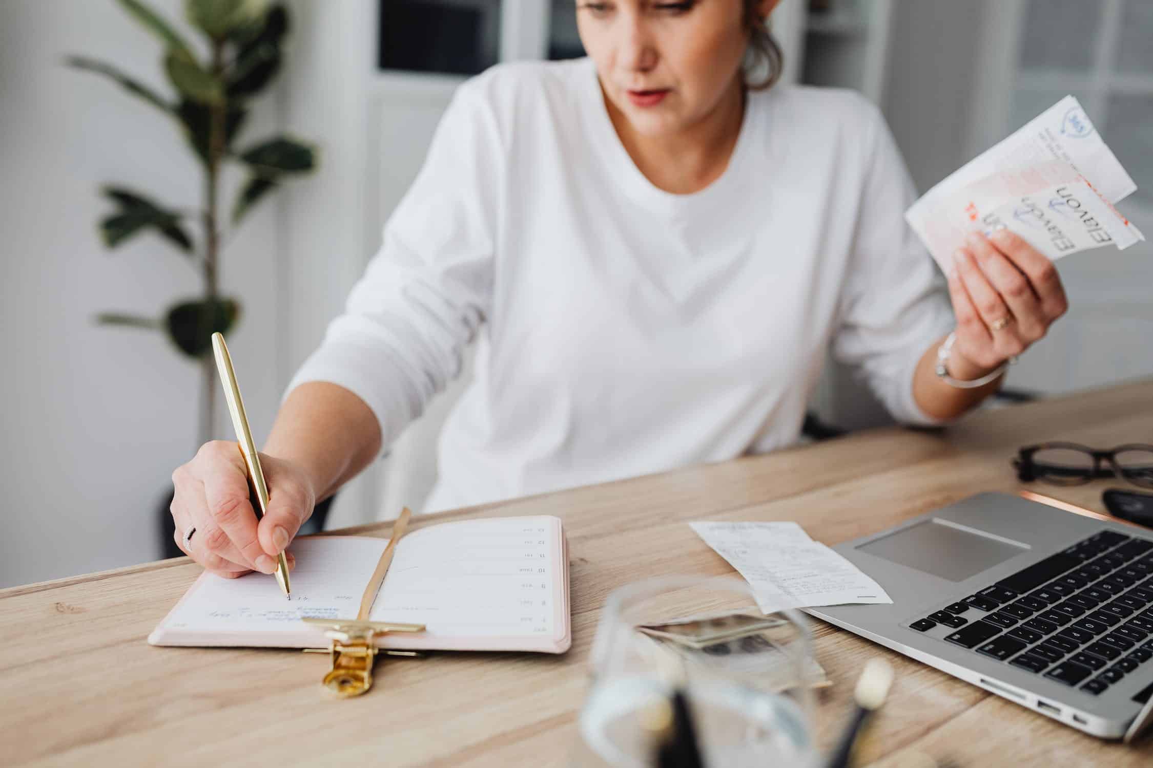 A person writes in a notebook while holding receipts, with a laptop and glass of water on a desk.