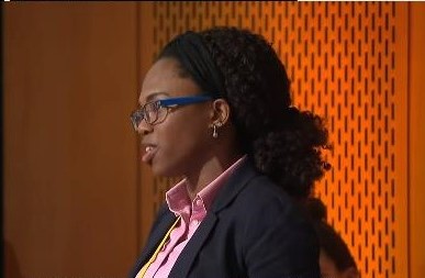 A woman with curly hair and glasses speaks during a presentation, wearing a blazer and a shirt, set against an orange patterned background.
