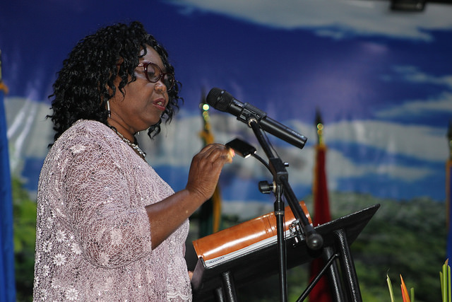 A woman with curly hair speaks at a podium, wearing a light-colored blouse, against a scenic background with flags and greenery.