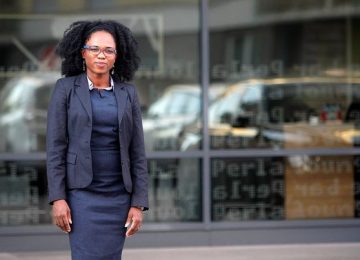 A woman in a business suit stands confidently outside a building, with glass windows and indistinct text in the background.