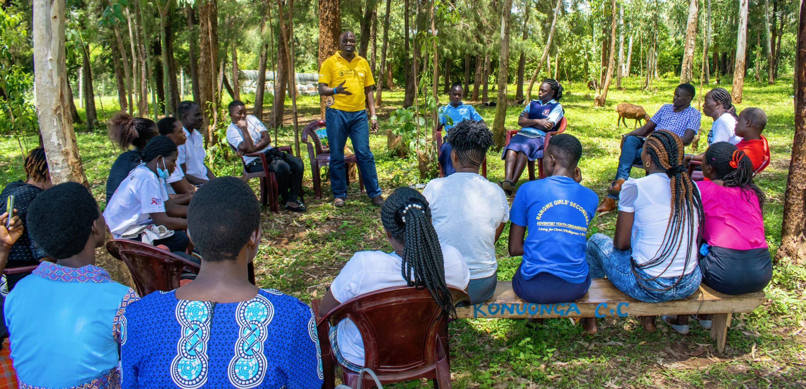 A group of people in a forested area engaged in a discussion, with one person addressing the gathering and others seated in a circle.